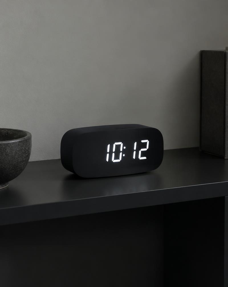 Black digital clock with white LED numerals on a dark shelf